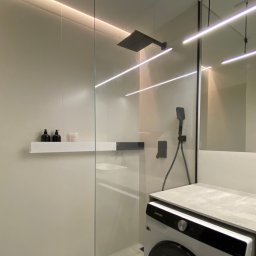Alfa Styl - Nowoczesna łazienka z kabiną prysznicową typu walk-in, oświetleniem LED i pralką w zabudowie. Minimalistyczny design z akcentami czerni.