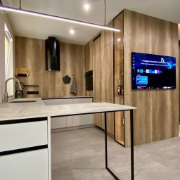 Alfa Styl - Nowoczesna kuchnia z wyspą i drewnianymi frontami szafek. Minimalistyczne oświetlenie LED, czarny okap i telewizor na ścianie. Szaro-biała kolorystyka.