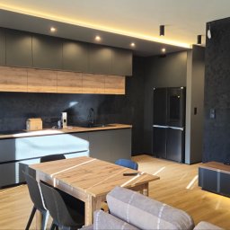 Alfa Styl - Nowoczesna aranżacja wnętrza: szara zabudowa kuchenna z drewnianymi akcentami, stół z krzesłami i czarna ściana z telewizorem. Ciepłe oświetlenie podkreśla detale.
