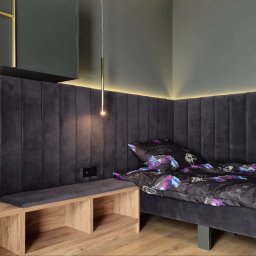 Alfa Styl - Nowoczesna sypialnia z panelem tapicerowanym za łóżkiem, podświetleniem LED i lampą wiszącą. Widoczna szafka nocna z drewnianym blatem i pościel z motywem kontrolerów do gier.