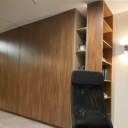 Alfa Styl - Nowoczesne biuro domowe: zabudowa z drewna z półkami, czarne krzesło biurowe, minimalistyczna lampa liniowa, szara ściana z oświetleniem punktowym i ciemne drzwi.