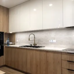 Nowoczesna kuchnia w odcieniach drewna i bieli. Uwagę przykuwa złota bateria i czarny zlew, a także zabudowana kuchenka mikrofalowa. Minimalistyczny design.