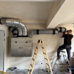 Greeno Sp. z o.o. - Montaż rekuperacji: instalator na drabinie łączy przewody wentylacyjne do centrali Regnet. Widoczne narzędzia i elementy instalacji. Ściany w stanie surowym.