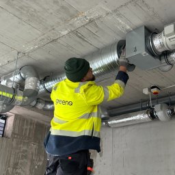 Greeno Sp. z o.o. - Pracownik w odblaskowej kurtce montuje system wentylacyjny z rurami na surowym betonowym suficie w budynku komercyjnym. Widoczne elementy instalacji HAVACo.