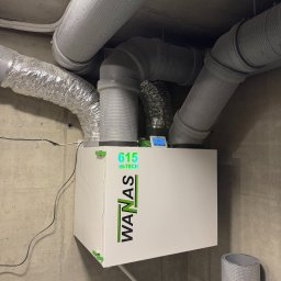 Greeno Sp. z o.o. - Biała jednostka rekuperacyjna WANAS 615 HI-TECH z podłączonymi przewodami wentylacyjnymi na tle surowego, betonowego sufitu i ściany. Widoczny wyświetlacz sterownika.