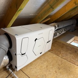 Greeno Sp. z o.o. - Biała centrala rekuperacyjna Thessla Green zainstalowana na poddaszu z widocznymi przewodami wentylacyjnymi i drewnianą konstrukcją dachu.