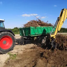 Żółta koparka CAT nabiera korzenie drzew i ziemię, ładując je do zielonej przyczepy ciągniętej przez zielony traktor Fendt z czerwonymi felgami na tle błękitnego nieba i zieleni drzew.