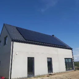 Nowoczesny dom jednorodzinny z szarym dachem i panelami fotowoltaicznymi zamontowanymi na połaci dachu, widoczne trzy okna i drzwi balkonowe, elewacja w kolorze surowego betonu, błękitne niebo.