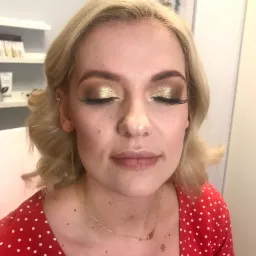 Portret kobiety z zamkniętymi oczami prezentującej złoty, błyszczący cień na powiekach i przedłużone rzęsy, delikatne fale blond włosów, czerwona bluzka w białe kropki i złoty łańcuszek z zawieszką.