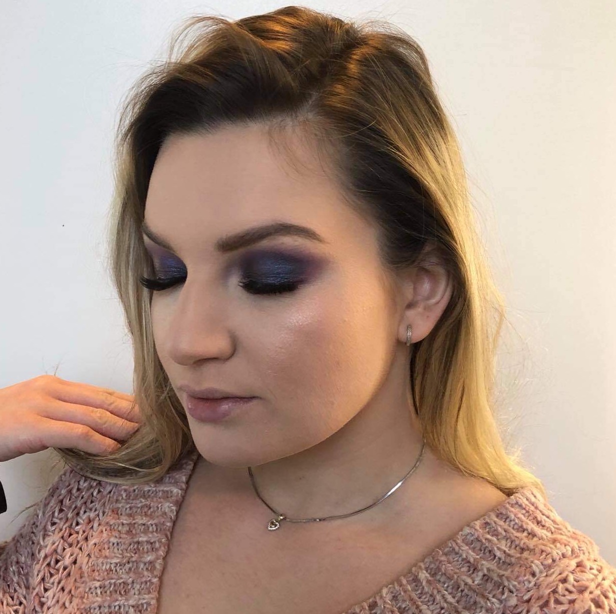 Portret kobiety z intensywnym, fioletowo-granatowym cieniowaniem powiek, długimi sztucznymi rzęsami i delikatnym błyszczykiem na ustach, podkreślone kości policzkowe, srebrny naszyjnik z zawieszką...