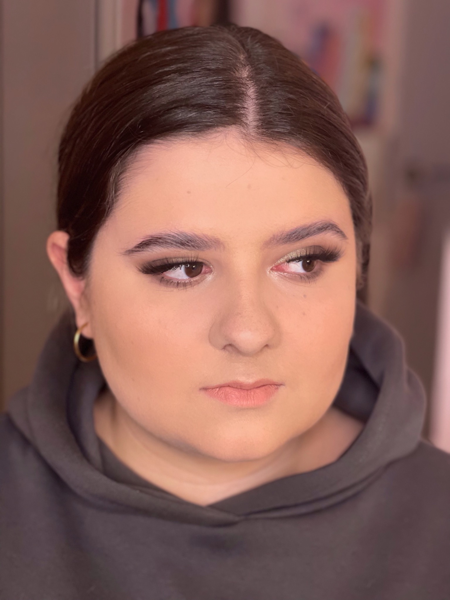 Portret kobiety z profesjonalnym makijażem, złoty cień na powiekach, wyraźnie podkreślone rzęsy, delikatny kontur ust, złote kolczyki koła, ubrana w szarą bluzę z kapturem, patrzy w bok.