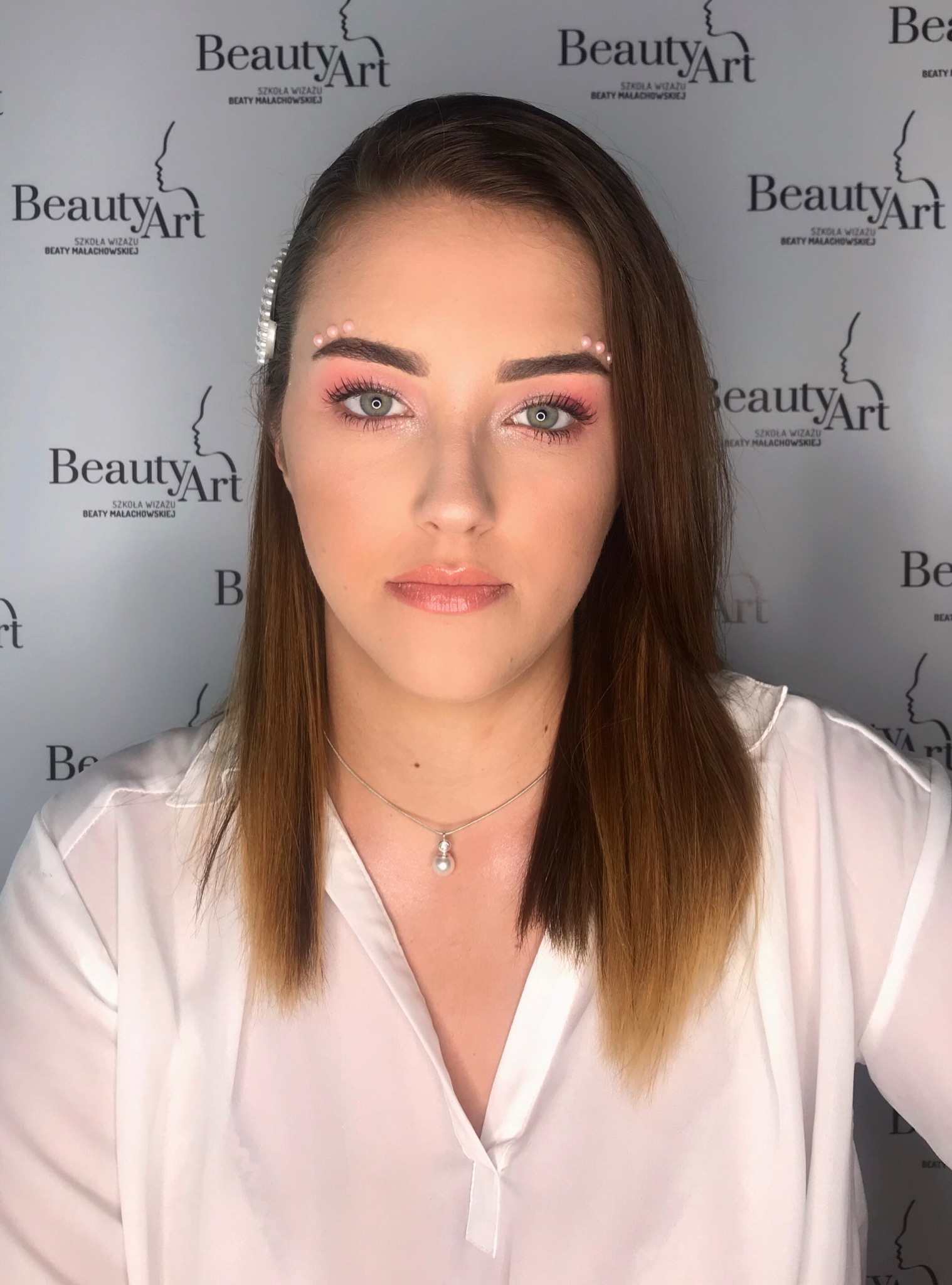 Portret kobiety z delikatnym, różowym makijażem oczu i ust, z ozdobnymi kropkami nad brwiami i perłową spinką we włosach, na tle z logo szkoły wizażu.
