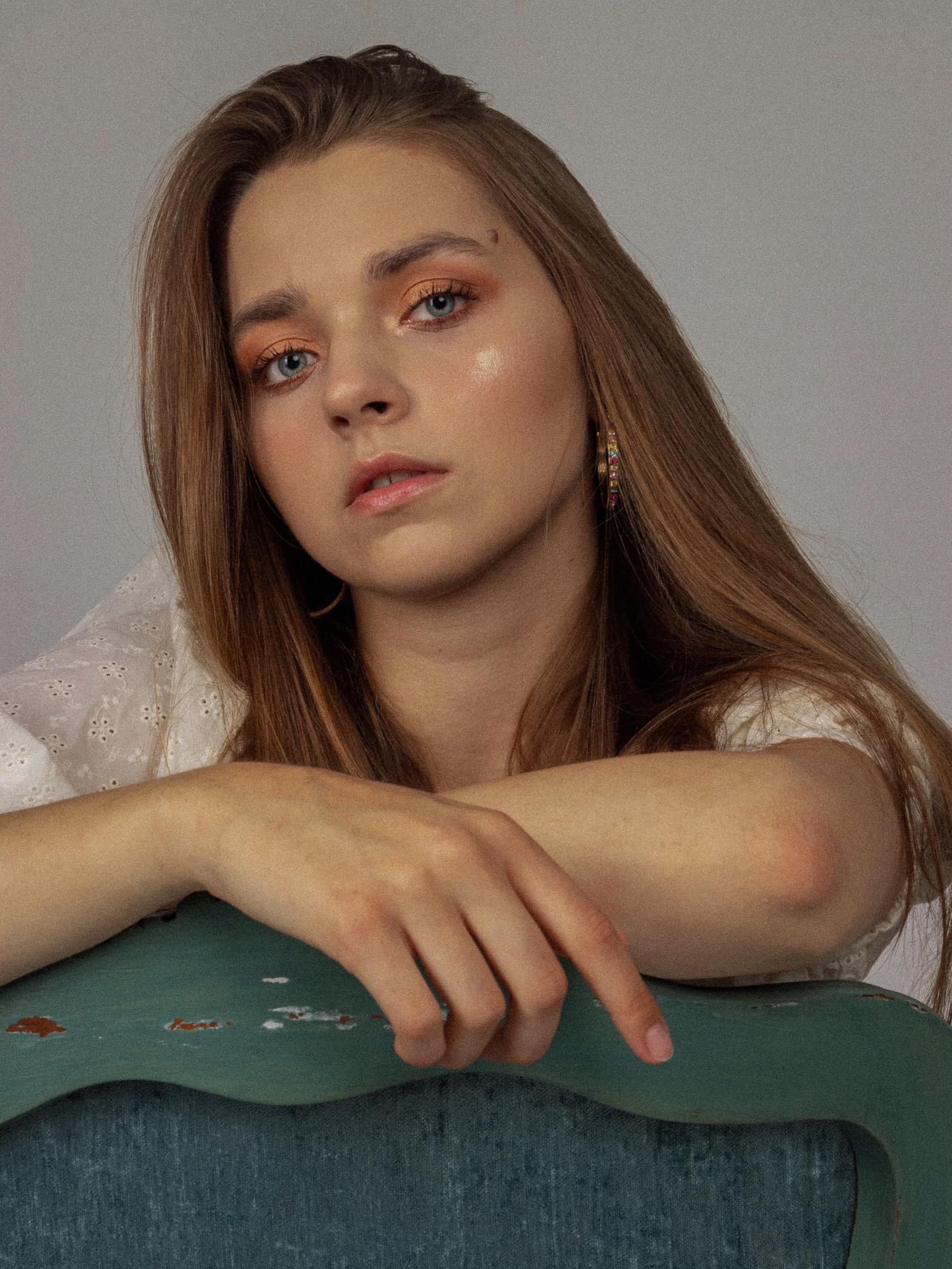 Portret młodej kobiety z pomarańczowym cieniem na powiekach, delikatnym błyszczykiem na ustach i złotymi kolczykami, opierającej ręce na zielonym krześle z przetarciami.