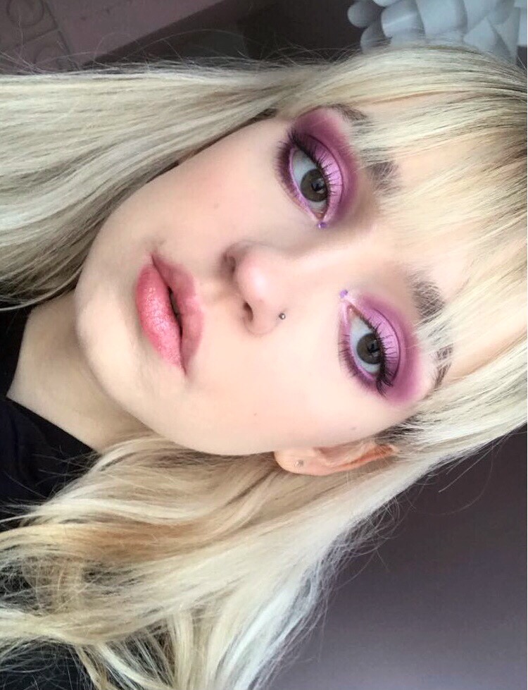 Portret kobiety z platynowymi włosami i intensywnym, fioletowym cieniowaniem powiek z błyszczącymi drobinkami, uzupełnionym o drobne, fioletowe naklejki pod oczami i delikatny róż na ustach.