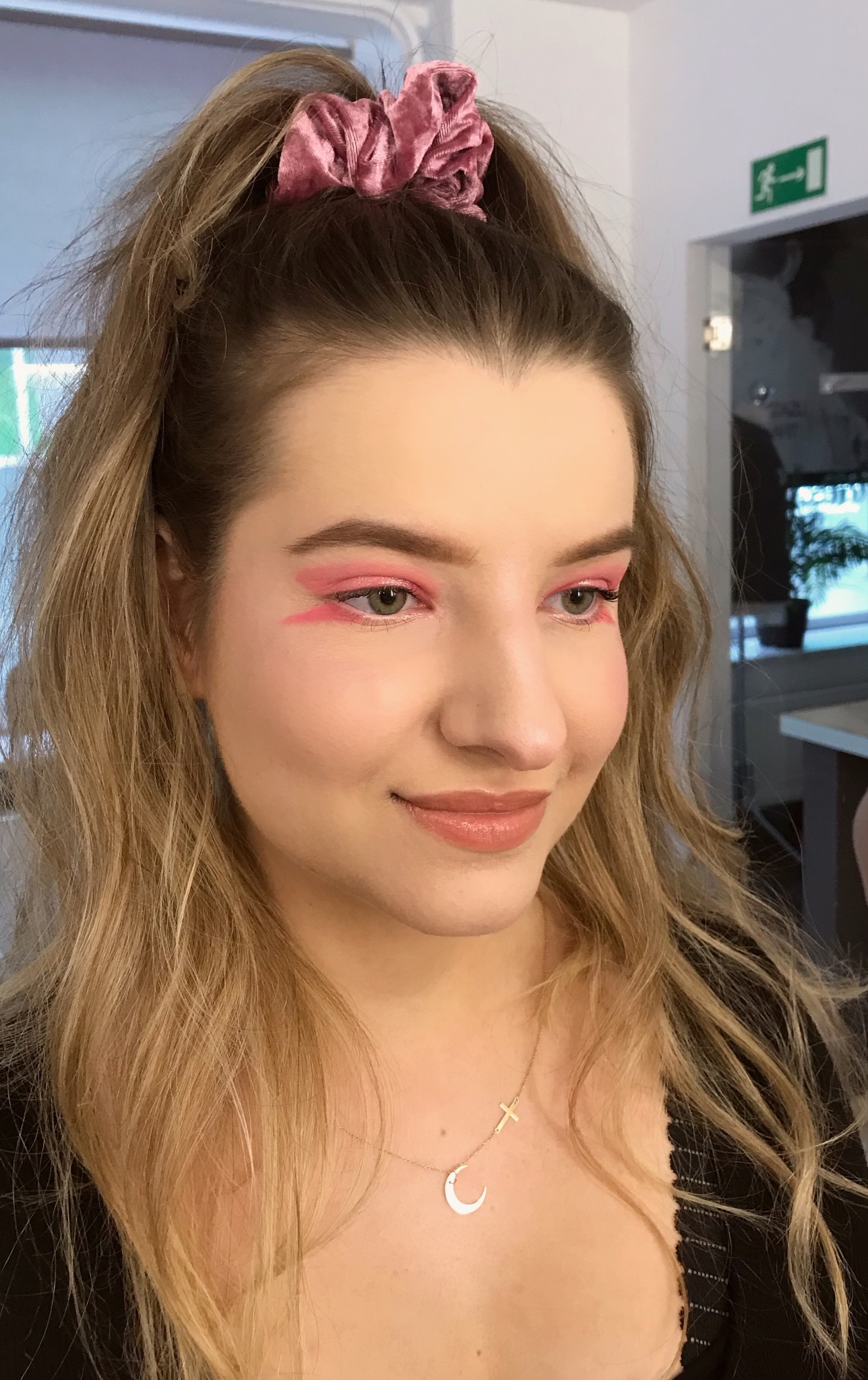 Portret uśmiechniętej młodej kobiety z różowym makijażem oczu w geometrycznym stylu, włosy upięte w wysoki kucyk z welurową gumką, delikatny naszyjnik z księżycem i krzyżykiem.