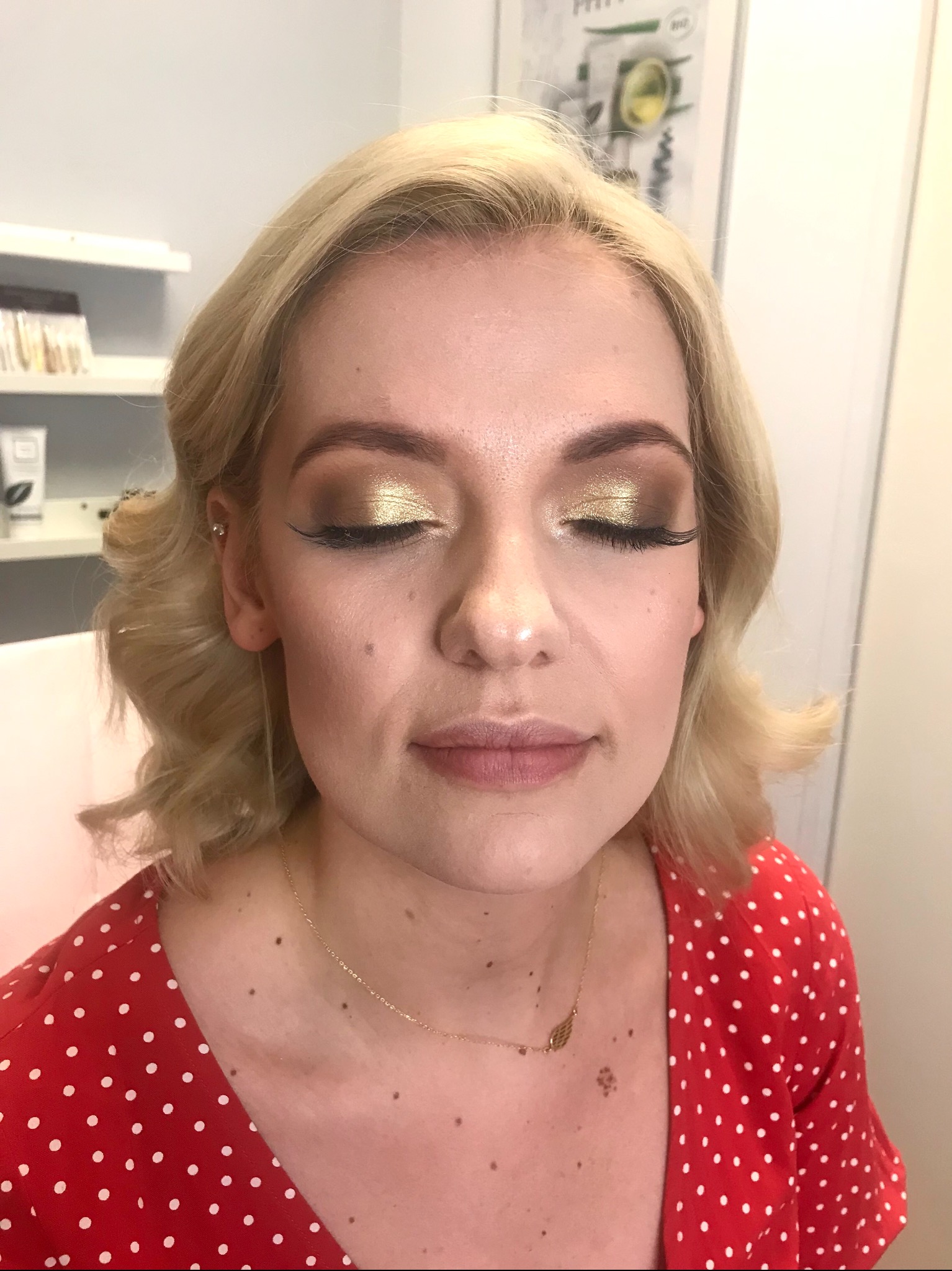 Portret kobiety z zamkniętymi oczami prezentującej złoty, błyszczący cień na powiekach i przedłużone rzęsy, delikatne fale blond włosów, czerwona bluzka w białe kropki i złoty łańcuszek z zawieszką.