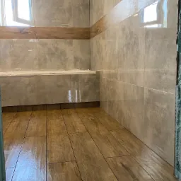 Wykończona łazienka z szarymi płytkami imitującymi beton na ścianach i drewnopodobną podłogą, widoczne otwarte okno i fragment zabezpieczonej folią wanny.