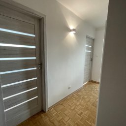 k DESIGNERS Przemysław Kufit - Korytarz po odświeżeniu z jasnymi ścianami, parkietem w jodełkę i dwiema parami drzwi z przeszkleniami w stylu skandynawskim, oświetlony kinkietem.