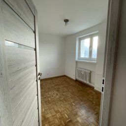 k DESIGNERS Przemysław Kufit - Pusty pokój z parkietem w jodełkę, białymi ścianami i grzejnikiem pod oknem, widoczny przez otwarte, szare drzwi z przeszkleniami.