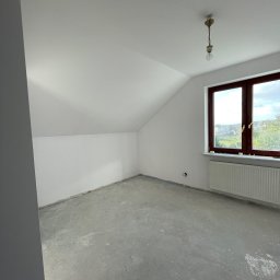 k DESIGNERS Przemysław Kufit - Pokój na poddaszu po malowaniu, z białymi ścianami i sufitem, szarym betonowym podłożem, grzejnikiem pod oknem i wiszącą lampą bez klosza.