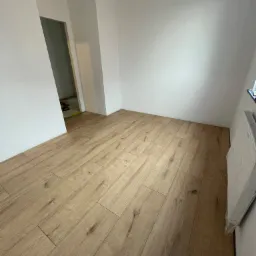 Jasne panele podłogowe ułożone w pustym, pomalowanym na biało pokoju, widok na próg drzwi do kolejnego pomieszczenia.