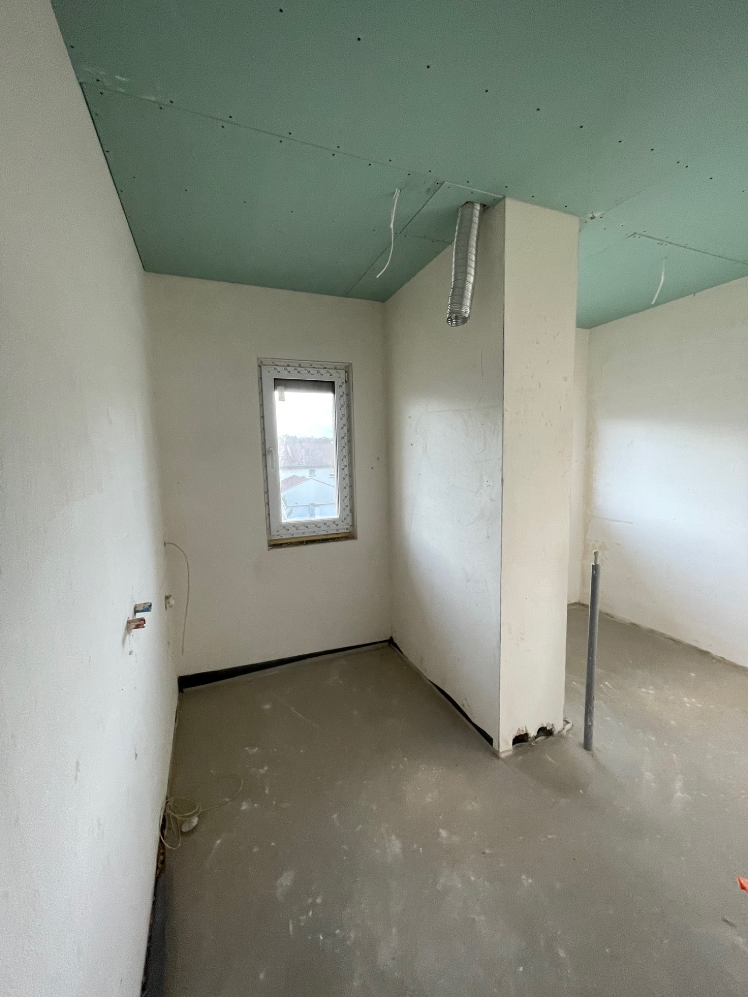 Surowe wnętrze w trakcie prac: zielona płyta na suficie, białe ściany z widocznymi instalacjami, wylewka na podłodze, aluminiowy przewód wentylacyjny.