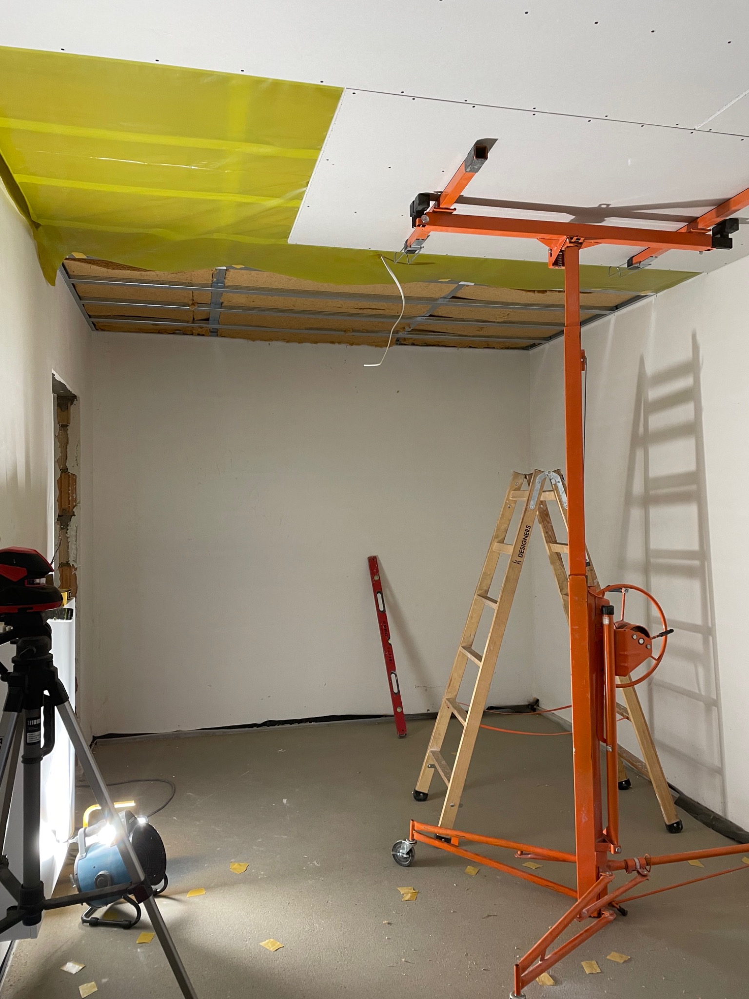 Montaż płyty na suficie za pomocą podnośnika, widoczna konstrukcja metalowa, żółta folia ochronna, poziomica laserowa na statywie i drabina.