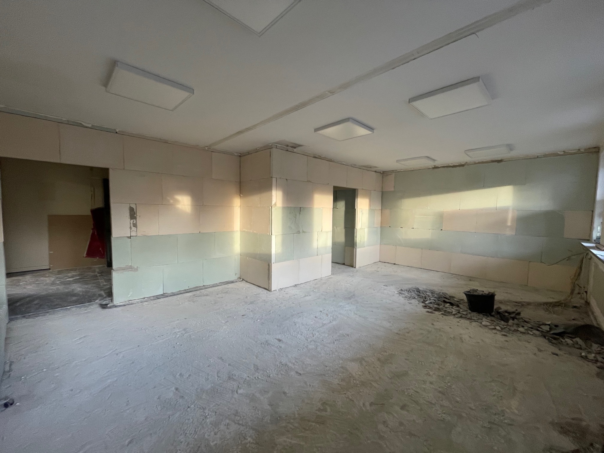 Wnętrze w trakcie prac przygotowawczych do wykończenia, widoczne ściany z bloków oraz ślady po wcześniejszych pracach na podłodze, oświetlenie sufitowe w kształcie prostokątów.