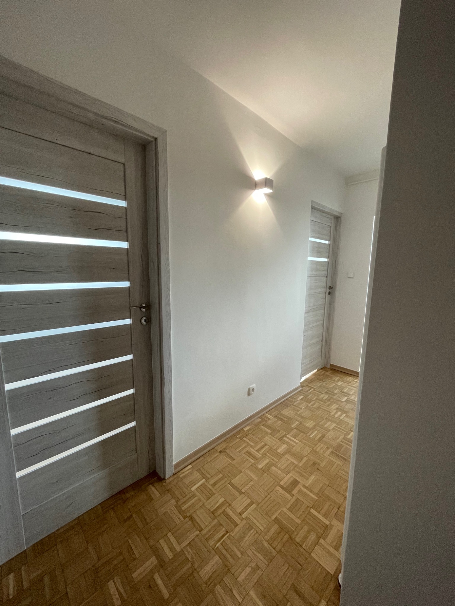 Korytarz po odświeżeniu z jasnymi ścianami, parkietem w jodełkę i dwiema parami drzwi z przeszkleniami w stylu skandynawskim, oświetlony kinkietem.