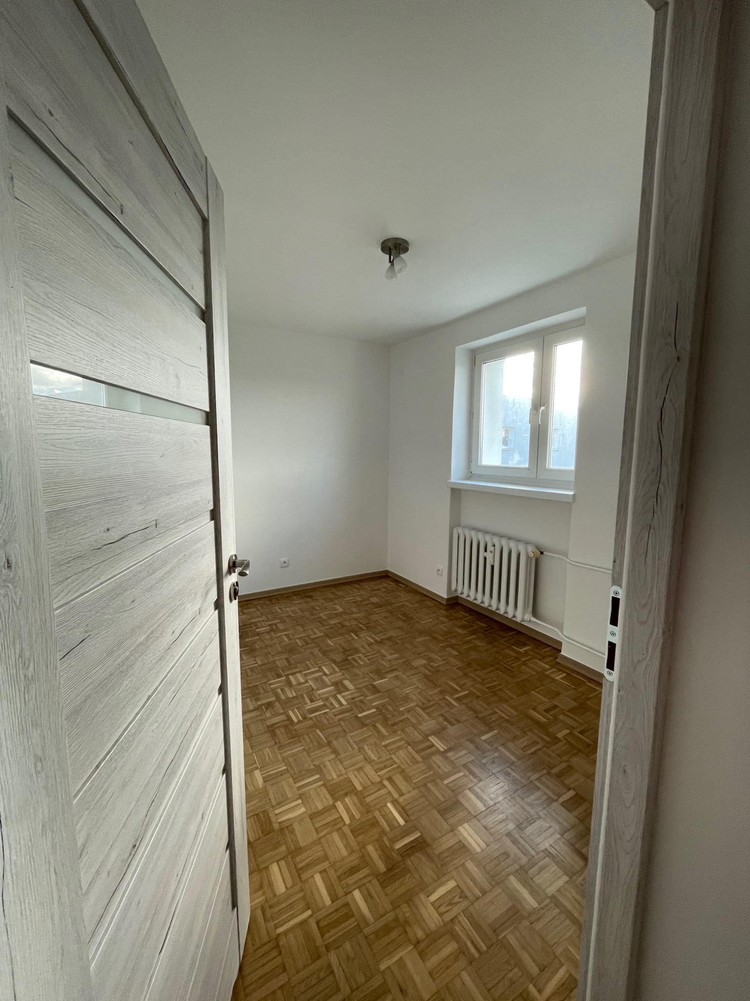 Pusty pokój z parkietem w jodełkę, białymi ścianami i grzejnikiem pod oknem, widoczny przez otwarte, szare drzwi z przeszkleniami.