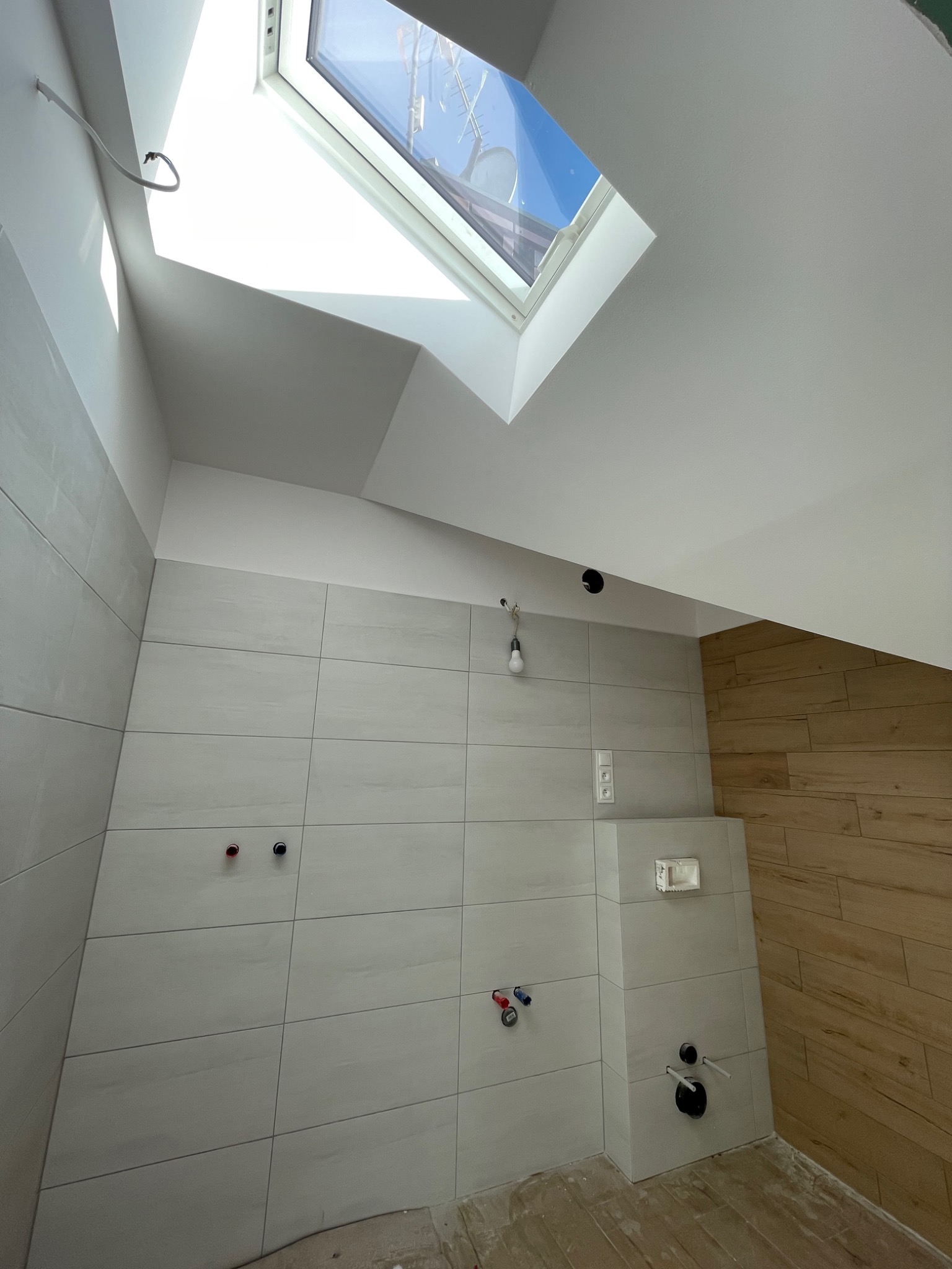 Wnętrze łazienki na poddaszu w trakcie wykańczania, widoczne płytki ceramiczne na ścianach, drewnopodobna okładzina, okno dachowe wpuszczające światło, oraz instalacja sanitarna i elektryczna...