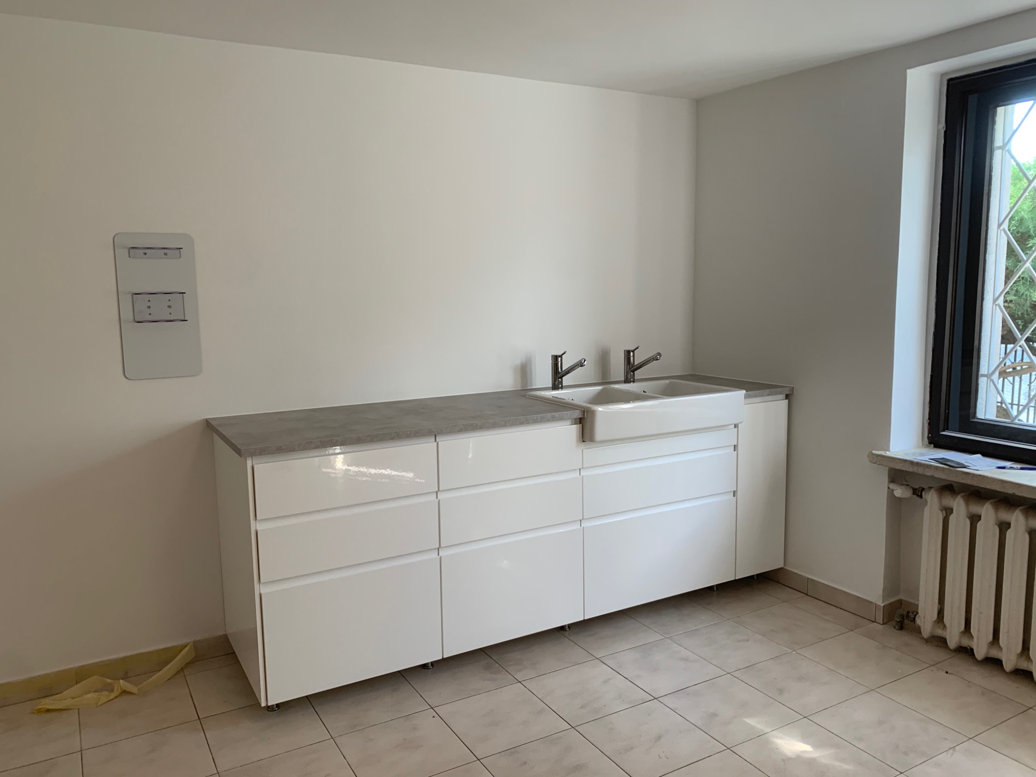 Jasne pomieszczenie po remoncie z białą zabudową meblową pod umywalki, szara posadzka z płytek, ściany w trakcie malowania, widoczny grzejnik pod oknem.