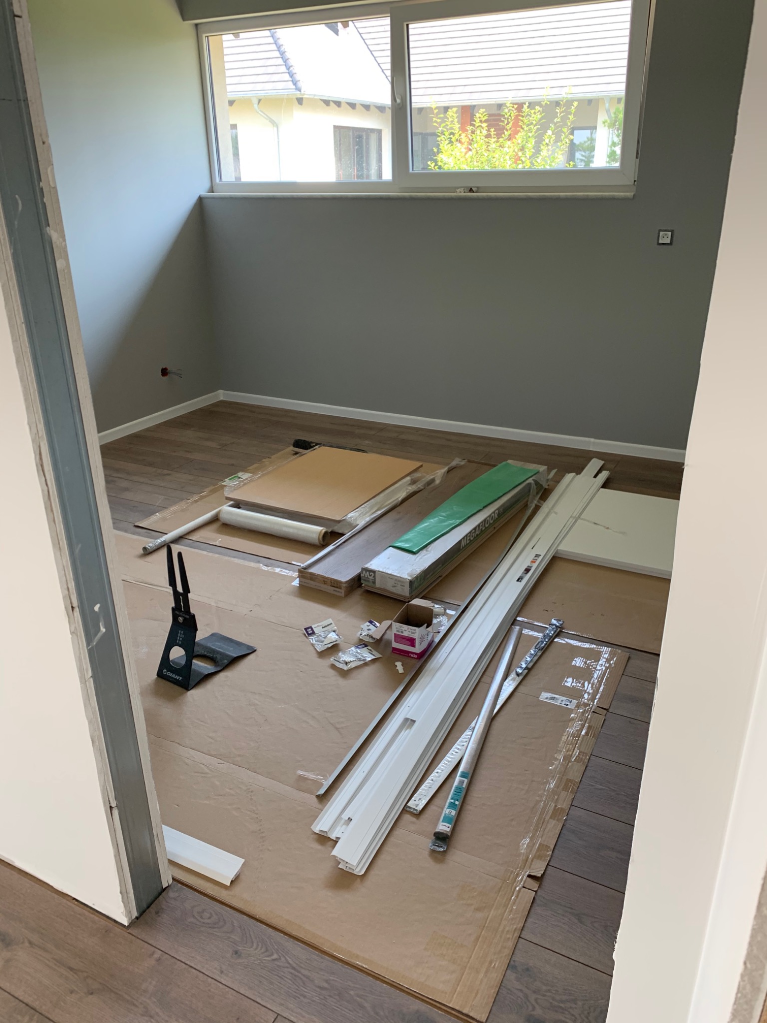 Przygotowania do montażu paneli podłogowych w pokoju z szarymi ścianami, widoczne narzędzia, listwy przypodłogowe, paczki z panelami oraz zabezpieczone podłoże kartonem.