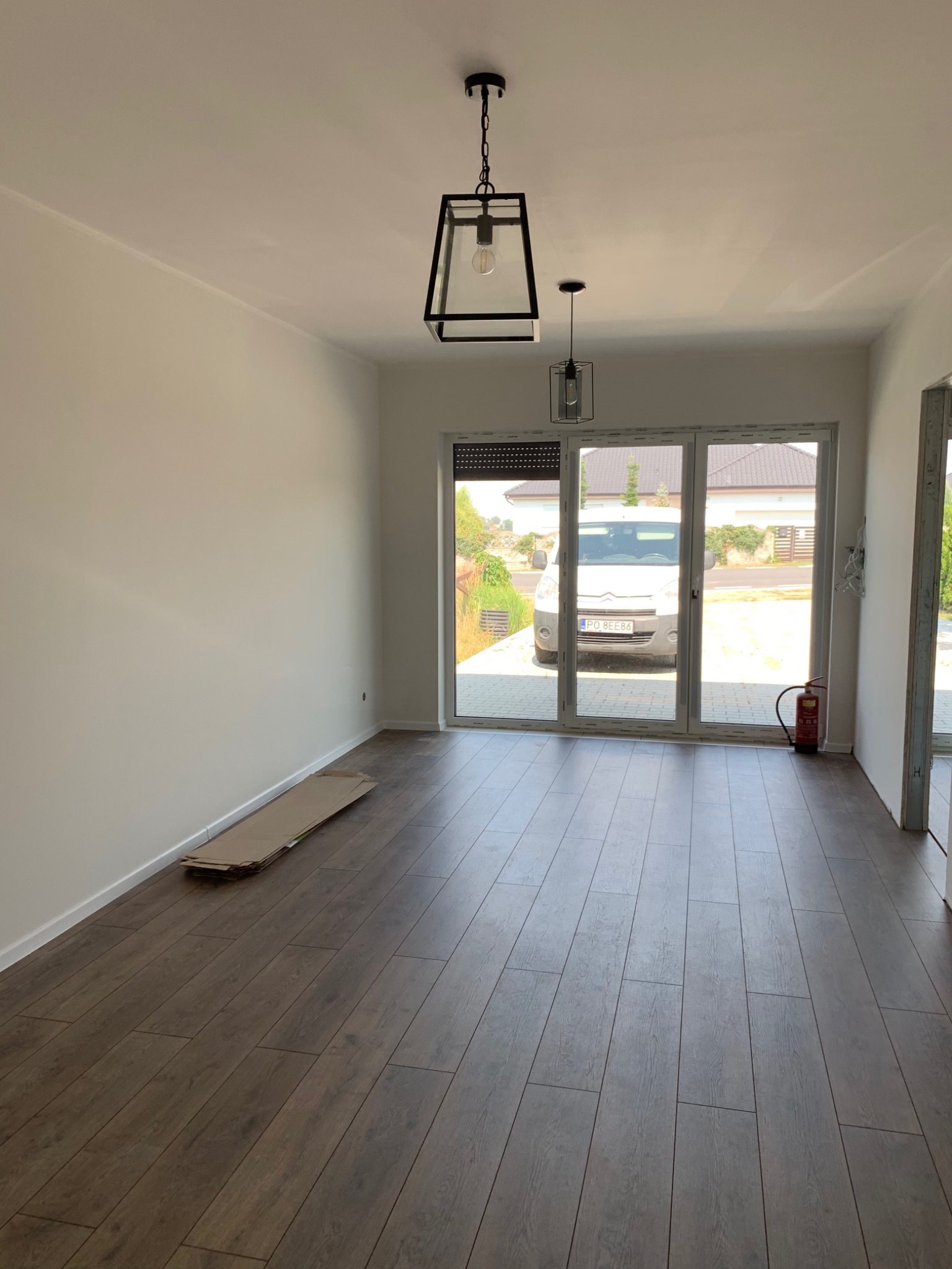 Wnętrze pokoju z nowymi panelami podłogowymi, świeżo pomalowanymi ścianami na biało, widoczne kartony na podłodze, okno balkonowe z widokiem na podjazd i samochód dostawczy oraz dwie nowoczesne...