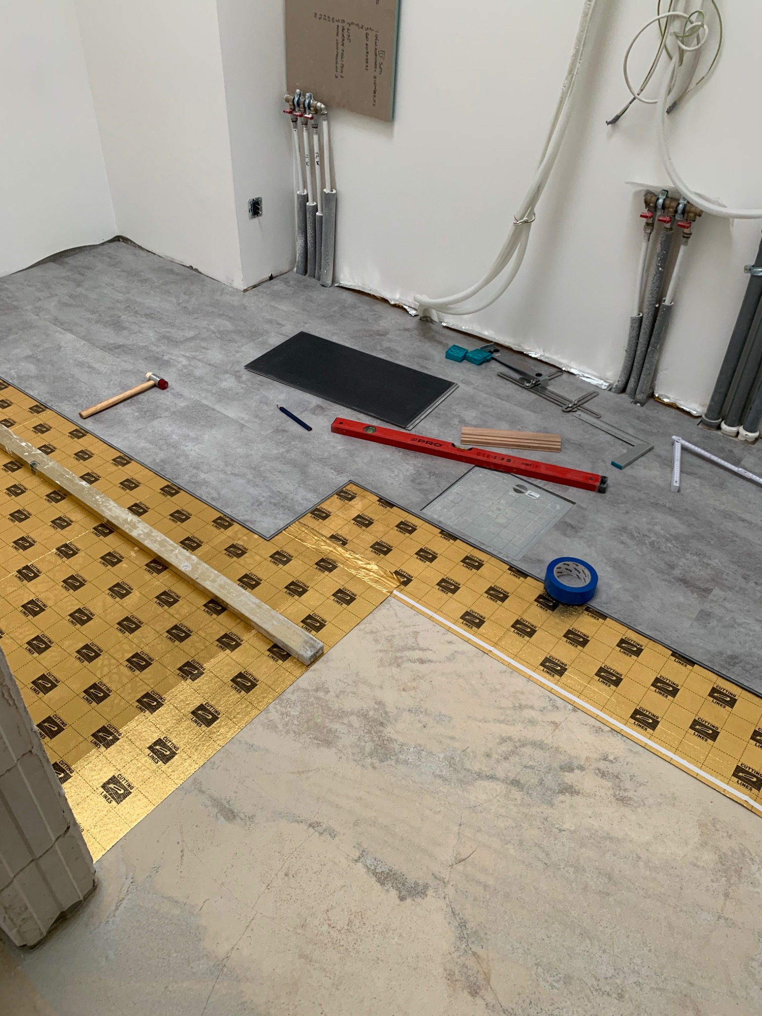 Układanie paneli podłogowych: widoczna folia podkładowa z nadrukiem, częściowo ułożone panele szare, narzędzia pomiarowe i wykończeniowe na podłodze, rury grzewcze przy ścianie.
