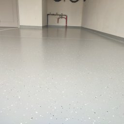 RAMI FLOOR RAFAŁ KLOCEK, MICHAŁ GORCZYCA SPÓŁKA CYWILNA - Jasnoszara posadzka z drobnymi, ciemnymi i jasnymi elementami w garażu. Widoczne rury instalacji na ścianie i listwy przypodłogowe.