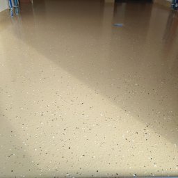 RAMI FLOOR RAFAŁ KLOCEK, MICHAŁ GORCZYCA SPÓŁKA CYWILNA - Beżowa posadzka z drobnymi, kolorowymi wtrąceniami, świeżo wylana, z widocznymi rurami instalacji przy ścianie. Gładka powierzchnia z refleksami światła.