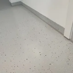 Jasnoszara posadzka z drobnymi, ciemnymi i białymi wtrąceniami, z listwą przypodłogową w podobnym wzorze. Widoczny fragment białych drzwi.