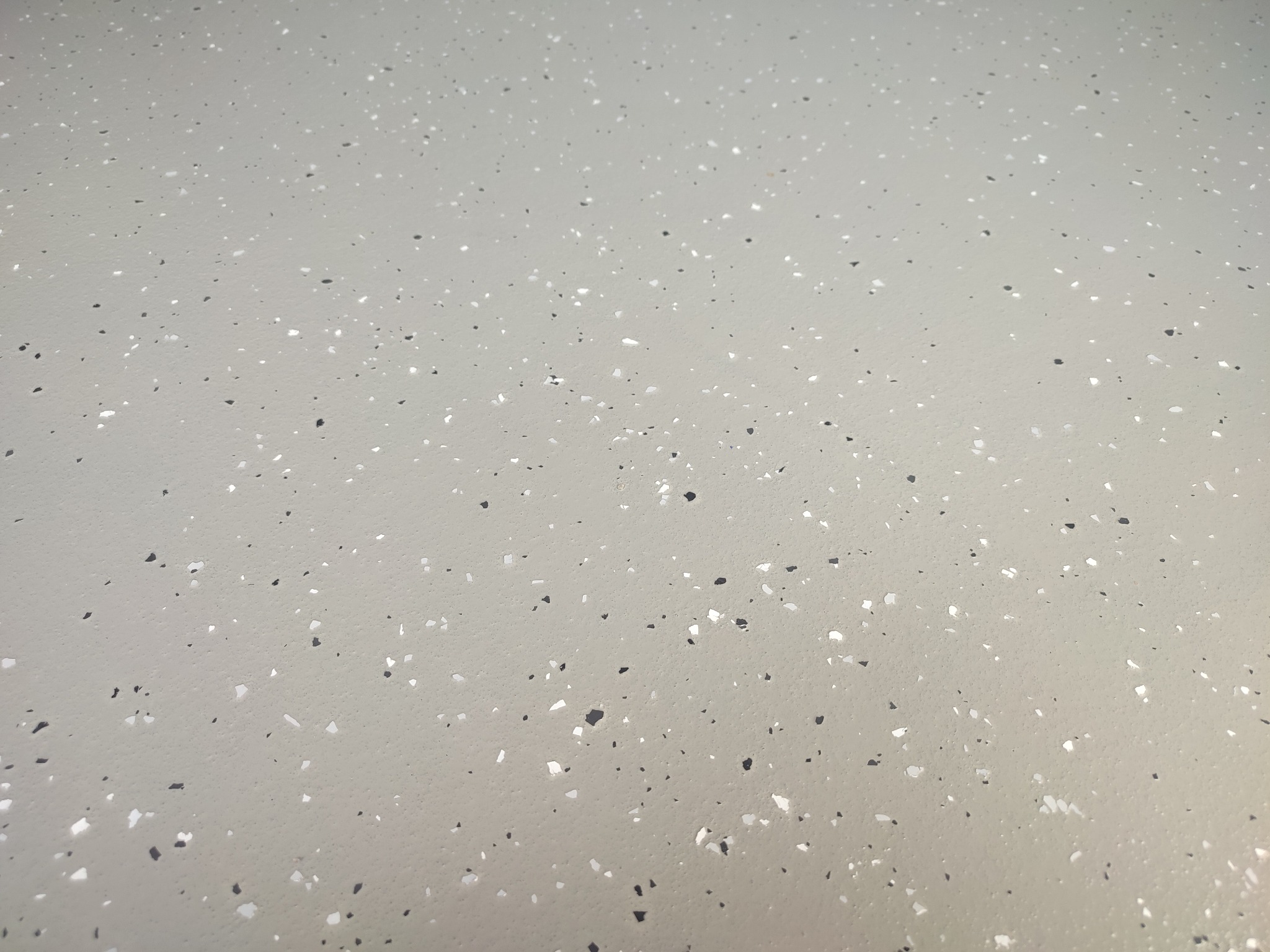 Jasnoszara posadzka żywiczna z drobnymi, czarnymi i białymi elementami dekoracyjnymi, tworzącymi wzór terrazzo. Gładka powierzchnia, równomierne rozłożenie wzoru.