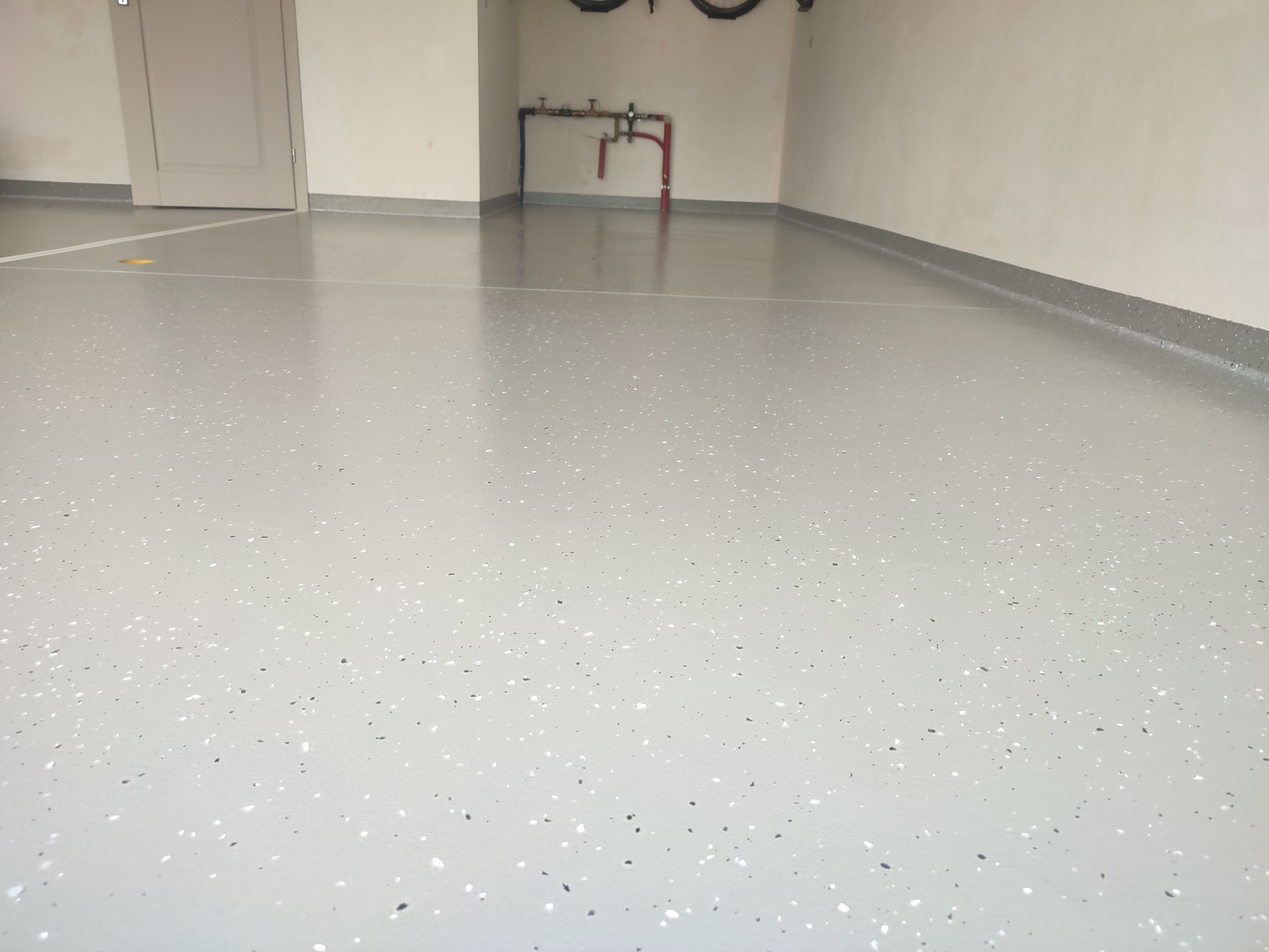 Jasnoszara posadzka z drobnymi, ciemnymi i jasnymi elementami w garażu. Widoczne rury instalacji na ścianie i listwy przypodłogowe.