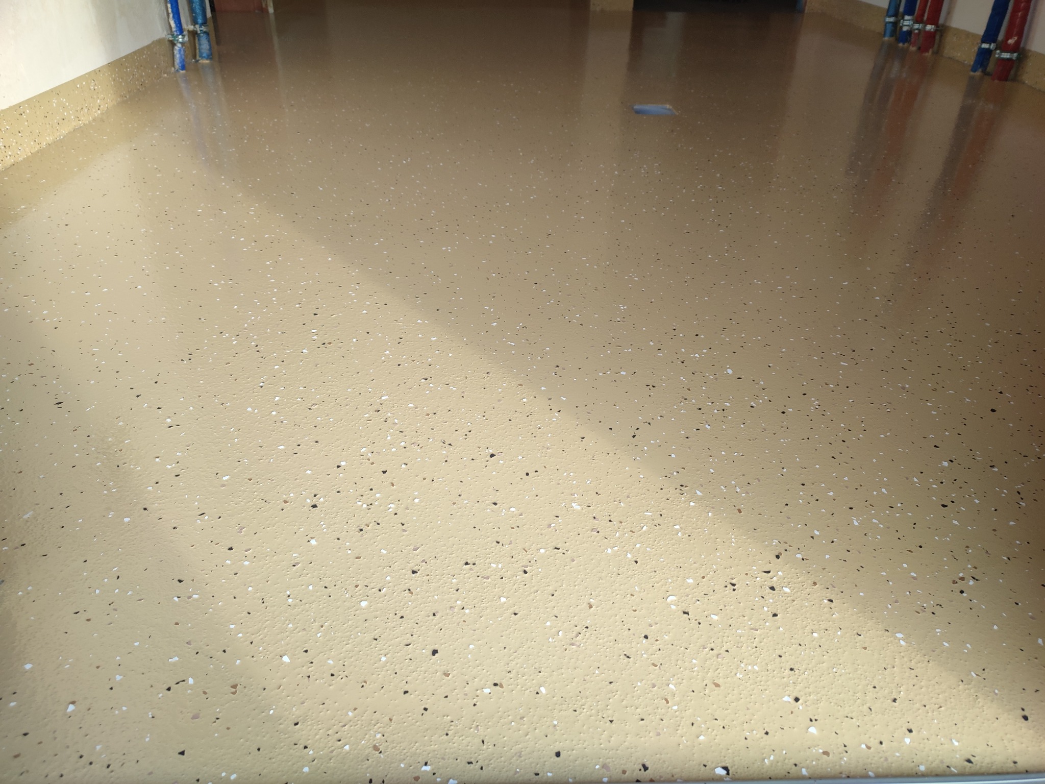 Beżowa posadzka z drobnymi, kolorowymi wtrąceniami, świeżo wylana, z widocznymi rurami instalacji przy ścianie. Gładka powierzchnia z refleksami światła.
