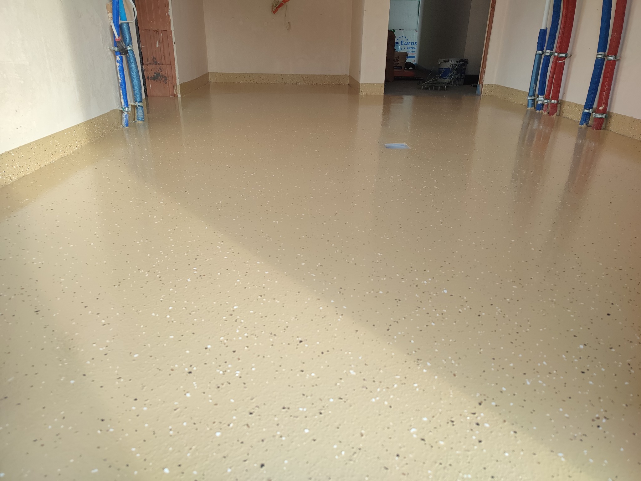 Beżowa, gładka posadzka z drobnymi, ciemnymi i jasnymi plamkami. Widoczne rury przy ścianie i wejście do innego pomieszczenia w tle.