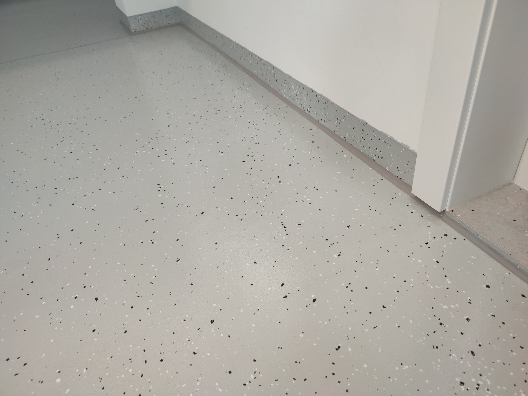 Jasnoszara posadzka z drobnymi, ciemnymi i białymi wtrąceniami, z listwą przypodłogową w podobnym wzorze. Widoczny fragment białych drzwi.