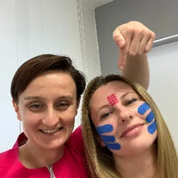 Dwie uśmiechnięte kobiety, jedna z aplikacjami z plastrów kinesiotaping na twarzy, wskazująca palcem na czoło z plastrem w kratkę.