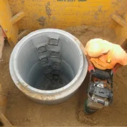 Pracownik w pomarańczowej kamizelce ubija ziemię wokół betonowej studni za pomocą zagęszczarki wibracyjnej, widoczna żółta obudowa wykopu.