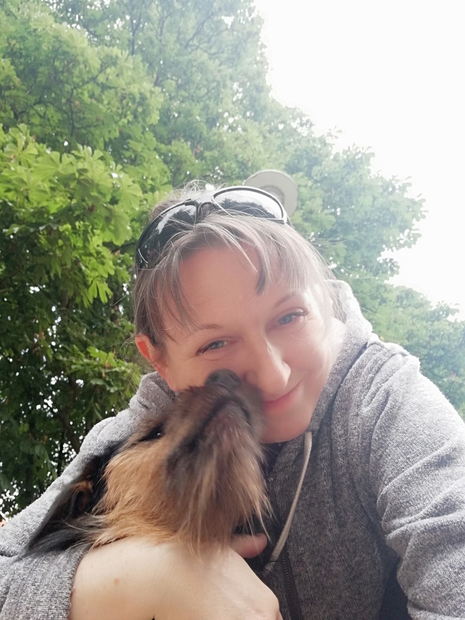 Uśmiechnięta kobieta w szarej bluzie z kapturem przytula brązowo-czarnego psa na tle zielonych drzew.