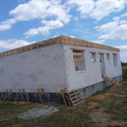 Surowy stan domu z białych bloczków, z widocznymi otworami okiennymi i drewnianym szalunkiem na dachu, wsparty tymczasowymi drewnianymi podporami, z paletą i opakowaniem bloczków Solbet obok.