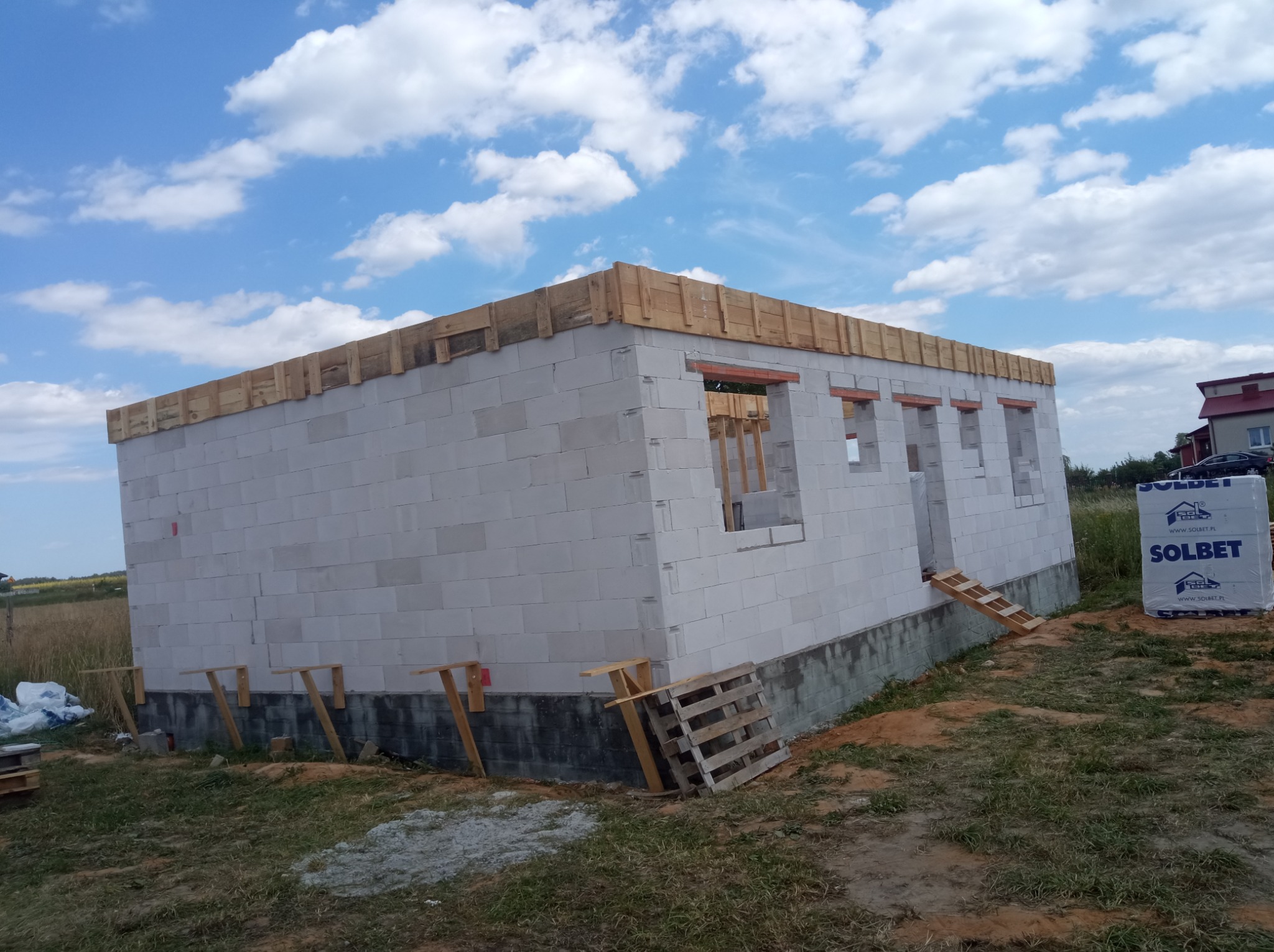 Surowy stan domu z białych bloczków, z widocznymi otworami okiennymi i drewnianym szalunkiem na dachu, wsparty tymczasowymi drewnianymi podporami, z paletą i opakowaniem bloczków Solbet obok.