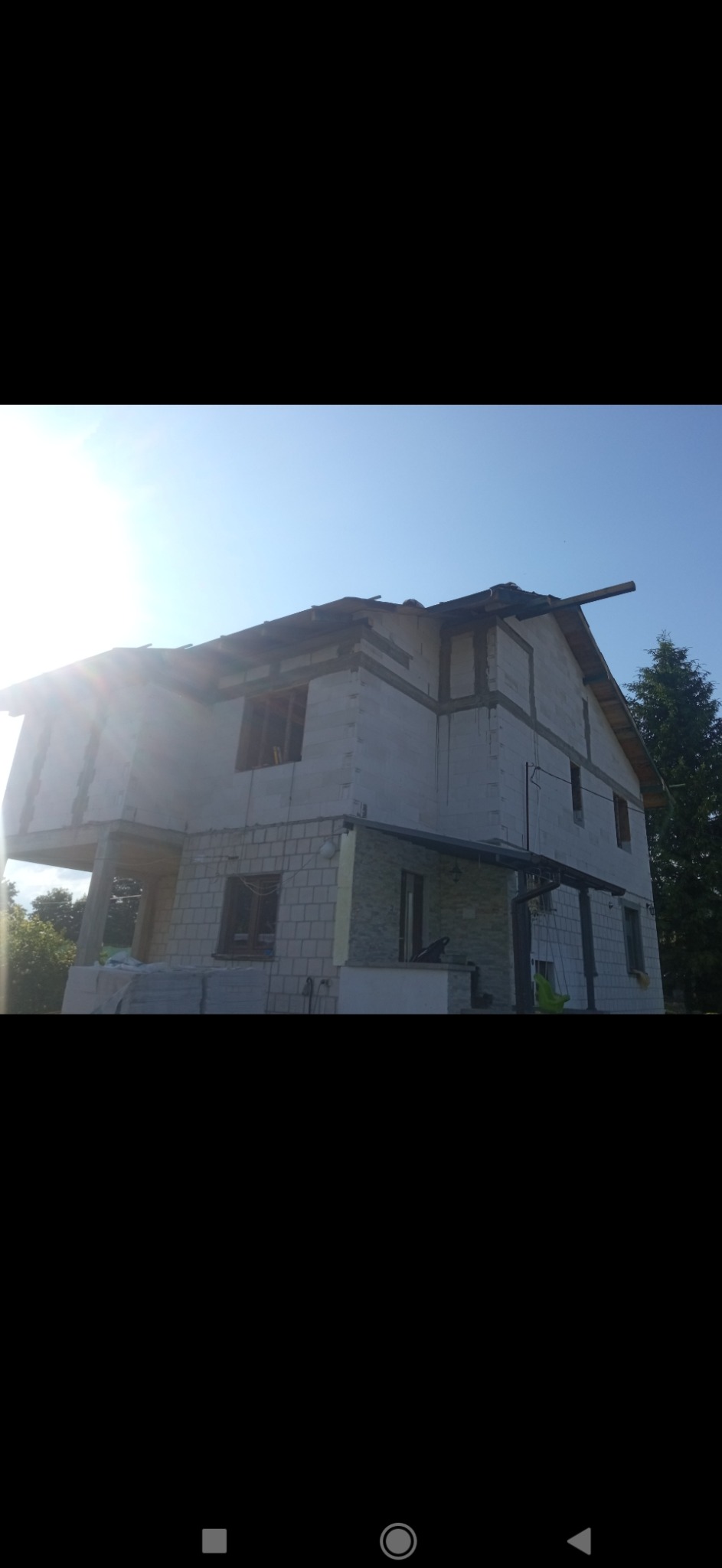 Dwupiętrowy dom w stanie surowym otwartym, z widocznymi ścianami z bloczków, częściowo osadzonymi oknami i elementami konstrukcji dachu, sfotografowany w słoneczny dzień.