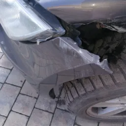 Uszkodzony przód samochodu: pęknięty zderzak, zarysowania i odsłonięte elementy konstrukcyjne przy kole. Widoczna opona zimowa i fragment felgi z logo BMW.